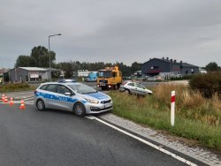 Ujęcie obrazujące miejsce zdarzenia uchwycone z większej odległości. Widoczny radiowóz oznakowany oraz uszkodzony Opel Corsa.