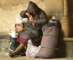 Bezdomna kobieta, siedząca na chodniku przy ścianie budynku. Obok niej wypełnione torby i foliowe reklamówki. Kobieta w trakcie posiłku - je jogurt.
