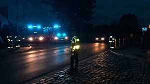 Sceneria nocna. Ulica w miejscowości Obra. Na pierwszym planie policjant kierujący ruchem w kamizelce odblaskowej. W tle inni funkcjonariusze w kamizelkach oraz nadające sygnały świetlne wozy straży pożarnej.