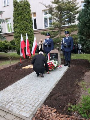Teren przed szkołą. Obelisk z tablicą pamiątkową poświęconą asp. Mieczysławowi Domagalskiemu - policjantowi zamordowanemu przez NKWD w 1940 roku. Pod pomnikiem wiązanka biało-czerwonych kwiatów składana przez jednego z gości.