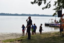 Policjant prowadzący działania rozpoczyna krótki wykład na temat bezpieczeństwa podczas wypoczynku nad wodą. W tle jezioro i pływające po nim jednostki.