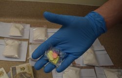 Policjant w rękawiczkach ochronnych pokazuje tabletki MDMA. Na drugim planie zabezpieczone dopalacze i banknoty.