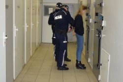 Policyjny areszt. Zatrzymana stoi twarzą do ściany na korytarzu aresztu. Dwaj policjanci zakładają na ręce zatrzymanej kobiety kajdanki.