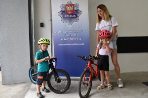 Mama z dwójką dzieci podczas akcji znakowania rowerów. Dzieci są wyposażone w rowerowe kaski ochronne. Wszyscy stoją na tle baneru KPP w Wolsztynie.