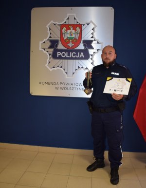 Policjant mł.asp. Mateusz Grelak eksponuje zdobyty medal oraz dyplom na tle gwiazdy Policji.