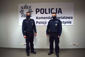 Policjanci w mundurach służbowych i maseczkach na tle ściany z napisem KOMENDA POWIATOWA POLICJI W WOLSZTYNIE