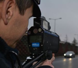 Zbliżenie na policjanta Wydziału Ruchu Drogowego podczas pomiaru prędkości miernikiem laserowym. Zdjęcie przedstawia fragment sylwetki policjanta oraz urządzenie którym się posługuje.