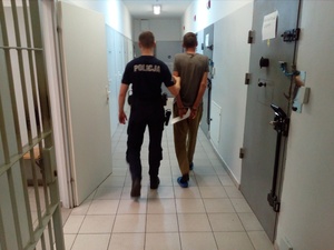 Policyjny areszt. Policjant doprowadza osadzonego, który ma założone kajdanki na rękach trzymanych z tyłu do pomieszczenia dla osób zatrzymanych.