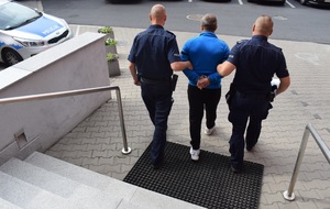 Schody przed wejściem głównym do budynku KPP w Wolsztynie. Dwaj umundurowani policjanci Wydziału Konwojowego prowadzą zatrzymanego do radiowozu, którym będą wykonywać konwój do miejsca, gdzie odbędzie karę pozbawienia wolności. Mężczyzna ubrany w niebieską bluzę dresową i ciemne spodnie znajduje się pomiędzy policjantami, którzy trzymają go za ręce spięte kajdankami z tyłu. Po lewej stronie widoczny fragment radiowozu oznakowanego.