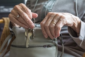 Zbliżenie na dłonie starszej kobiety o wyraźnie pomarszczonej skórze. Kobieta trzyma w dłoniach pęk kluczy do zamków w drzwiach. Dłonie opiera o szarą damską torebkę.