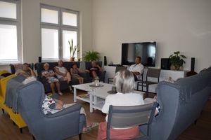 Pomieszczenie Domu Dziennego Pobytu Senior+ w Wolsztynie. Na kanapach i fotelach ustawionych w literę C siedzą seniorzy naprzeciw nich siedzący na krześle policjant Wydziału Kryminalnego podczas prelekcji.