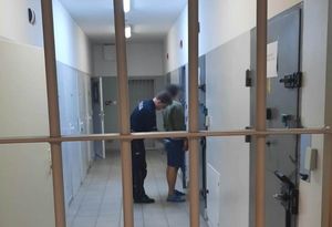 Areszt. Widok przez zamknięte kraty. Policjant pochyla się nad stojącym przy ścianie osadzonym i przeszukuje go przed przystąpieniem do przesłuchania.