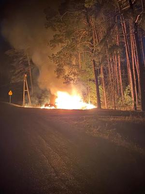 Sceneria nocna. Las. Na poboczu drogi słup ognia powstały w wyniku pożaru samochodu. Na drugim planie widoczne drzewa i unoszący się dym.