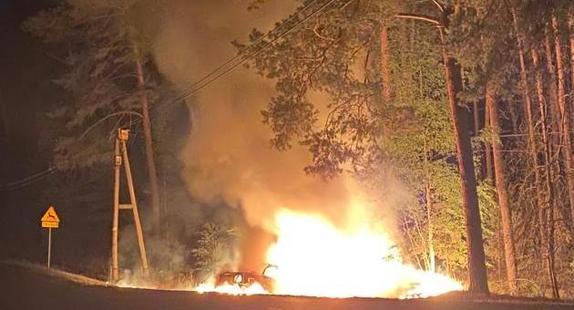 Sceneria nocna. Las. Na poboczu słup ognia powstały w wyniku pożaru samochodu. Na drugim planie widoczne drzewa i unoszący się dym.