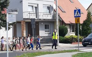 Akcja &quot;Pomachaj kierowcy&quot;. Policjantka podczas zajęć praktycznych z dziećmi przechodzi przez oznakowane przejście dla pieszych przy Szkole Podstawowej nr 5 w Wolsztynie. Dzieci machają do kierowcy samochodu osobowego, który zatrzymał się przed pasami.