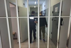 Na pierwszym planie widoczne kraty koloru szarego, za którymi znajdują się idący korytarzem policjant i osadzony w areszcie mężczyzna. Policjant trzyma go za ramię i doprowadza do pomieszczenia w którym przebywa podczas zatrzymania.