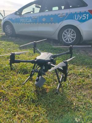 Teren trawiasty. Na pierwszym planie dron stojący na trawniku, za nim oznakowany radiowóz.