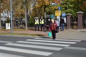 Policjanci Wydziału Ruchu Drogowego podczas obserwacji ruchu pieszych przed wejściem głównym na cmentarz. Na przejściu widoczna starsza kobieta przechodząca na drugą stronę ulicy.