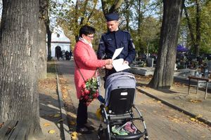 Cmentarz. Dzielnicowy rozmawia z kobietą z wózkiem. Przekazuje jej ulotkę w której zawarte są zasady bezpieczeństwa podczas odwiedzania cmentarzy.