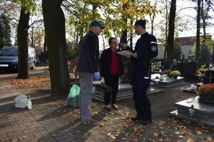 Cmentarz. Dzielnicowy rozmawia z kobietą i mężczyzną porządkującymi grób. Przekazuje im ulotkę w której zawarte są zasady bezpieczeństwa podczas odwiedzania cmentarzy.