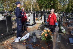Cmentarz. Dzielnicowy rozmawia z małżeństwem, które sprząta grób.  Przekazuje ulotkę w której zawarte są zasady bezpieczeństwa podczas odwiedzania cmentarzy.