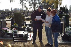 Cmentarz. Dzielnicowy rozmawia z dwiema kobietami, które sprzątają grób.  Przekazuje im ulotkę w której zawarte są zasady bezpieczeństwa podczas odwiedzania cmentarzy.