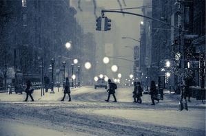 Sceneria zimowa. Szeroka ulica dużego miasta podczas śnieżycy. Ludzie idąc jeden za drugim przechodzą na drugą stronę ulicy. W tle latarnie miejskie oraz zawieszona nad jezdnią sygnalizacja świetlna.
