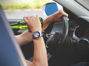 Zdjęcie ilustracyjne. Fragment postaci siedzącej wewnątrz samochodu. Jedna ręka spoczywa na kierownicy, w drugiej znajduje się przechylona w sposób sugerujący picie butelka alkoholu.
