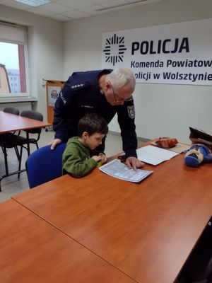 Chłopiec siedząc przy stole odczytuje swoje nazwisko na okolicznościowym adresie na pamiątkę wizyty w KPP w Wolsztynie. Nad dzieckiem pochyla się policjant.