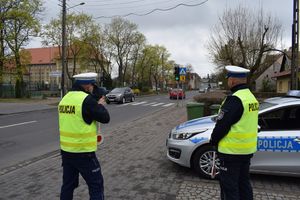 Dwaj policjanci służby ruchu drogowego stoją obok zaparkowanego na ulicy Poniatowskiego w Wolsztynie radiowozu kontrolując ruch pojazdów. Jeden z policjantów mierzy prędkość poruszających się pojazdów, drugi stoi obok niego.