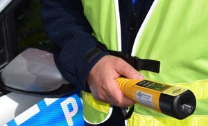 Zbliżenie na policjanta stojącego przy radiowozie oznakowanym i trzymającego w dłoni alkotest.