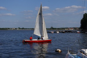 Czerwona żaglówka z białym żaglem na wodzie jeziora nieopodal przystani. W żaglówce znajdują się 3 osoby. Na drugim tle widoczne są pomosty przystani.