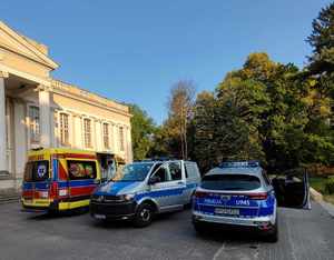 Pojazdy służbowe policji oraz Służby Zdrowia stojące przed pałacem w parku w Wolsztynie. Policjanci i ratownicy medyczni znajdują się poza pojazdami, na brzegu jeziora prowadząc akcję ratunkową.