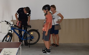 Policjant podczas znakowania roweru. Obok stoi rodzina - mama i dwóch synów przypatrując się czynności. Chłopcy mają założone na głowach rowerowe kaski ochronne.