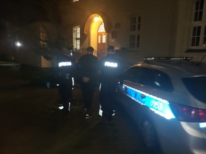 Sceneria nocna. Zaparkowany przed budynkiem sądu radiowóz oznakowany. Policjanci prowadzą zatrzymanego na salę rozpraw.