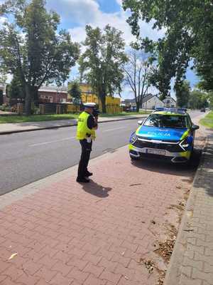 Stojący przy radiowozie policjant mierzy prędkość pojazdów na drodze