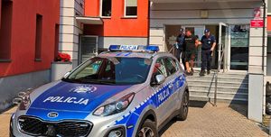 Wejście główne budynku KPP w Wolsztynie. Dwoje policjantów idąc w dół po schodach prowadzą zatrzymanego mężczyznę w kierunku radiowozu. Mężczyzna trzymany jest za ręce.