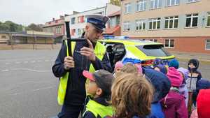 Policjant stoi przed grupą dzieci na boisku szkolnym prezentuja pałkę wielofunkcyjną TONFA. Na drugim plani budynek szkoły.