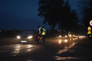 Sceneria nocna. Rząd stojących a drodze pojazdów. Policjanci w żółtych kamizelkach kontrolują trzeźwość kierowców.