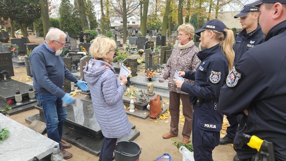 Policjanci rozmawiają na cmentarzu z ludźmi porządkującymi groby oraz wręczają im ulotki z poradami dotyczącymi bezpieczeństwa.