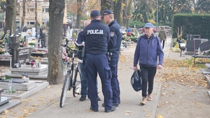 Policjanci rozmawiają na cmentarzu z ludźmi porządkującymi groby oraz wręczają im ulotki z poradami dotyczącymi bezpieczeństwa.