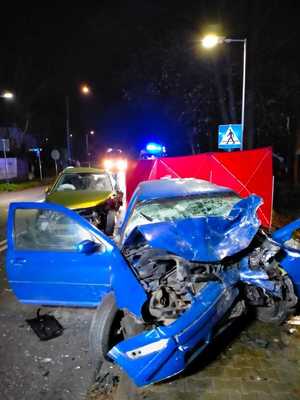 Sceneria nocna dwa rozbite pojazdy zajmujące część jezdni. Obok policjanci i strażacy podczas czynności na miejscu wypadku.