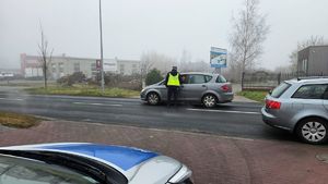 Sceneria dzienna, lekkie zamglenie. Policjant ubrany w żółtą kamizelkę odblaskową kontroluje trzeźwość kierujących pojazdami.