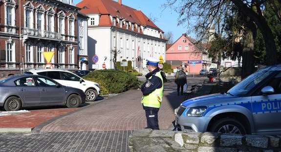 Umundurowany policjant stojąc obok radiowozu obserwuje ruch uliczny.