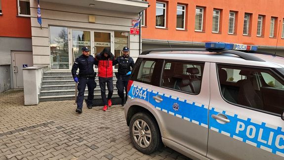 Dwaj umundurowani policjanci doprowadzają zatrzymanego do rafiowozu.