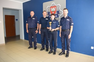 Zdjęcie grupowe na tle granatowej ściany. Policjantka trzymająca puchar i trzech policjantów stojących po obu jej stronach.