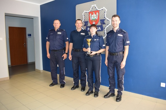 Zdjęcie grupowe na tle granatowej ściany. Policjantka trzymająca puchar i trzech policjantów stojących po obu jej stronach.