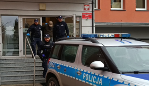Na zewnątrz - teren przy głównym wejściu do komendy. Policjanci doprowadzają zatrzymanego do radiowozu