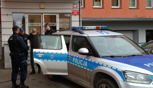 Na zewnątrz - teren przy głównym wejściu do komendy. Policjanci doprowadzają zatrzymanego do radiowozu