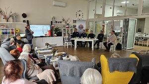 Debata społeczna w Domu Dziennym Senior+. Prelegent stojąc przy ekranie o dużej przekątnej referuje jeden z tematów, uczestnicy siedzą na fotelach, a organizatorzy za stołem prezydialnym.